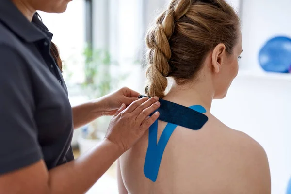 Chinesische Massage-Therapeutin klebt Kinesio-Tape auf die Schultern und den Hals einer attraktiven blonden Kundin in einer hellen Arztpraxis — Stockfoto