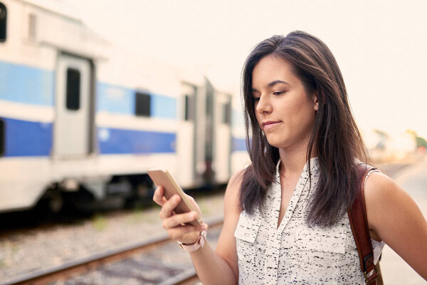 Confident millennial student on the go checking her smart phone on a train platform