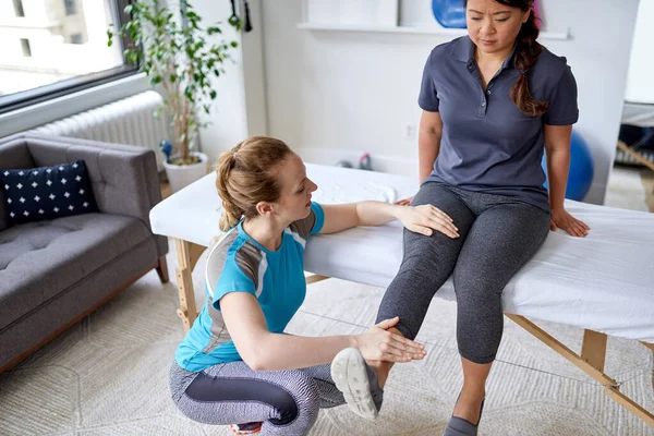 Fisioterapeuta mujer caucásica estirando la pierna y la rodilla de una paciente china de mediana edad sentada en una mesa de masaje — Foto de Stock