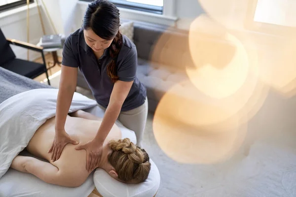 Mulher chinesa massagem terapeuta dando um tratamento para um cliente loiro atraente na mesa de massagem em um escritório médico brilhante — Fotografia de Stock