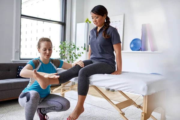 Белая женщина-физиотерапевт, растягивающая ногу и колено взрослой китайской пациентки, сидящей на массажном столе — стоковое фото