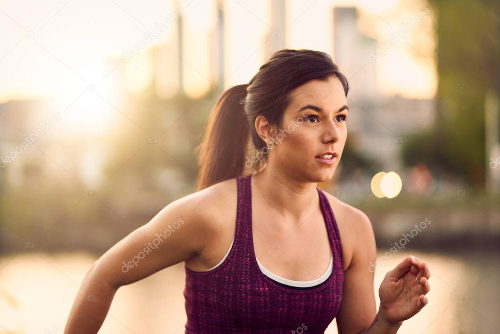 Portrait of active millenial woman jogging at dusk with an urban cityscape and sunset in the background