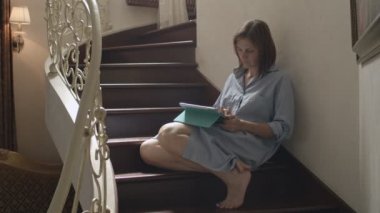 Genç esmer kadınlar ahşap merdivenlerde oturan ve tablet üzerinde çalışıyor. Klasik iç tablet ile konuşan kadın yetişkin