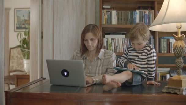 Blond toddler boy disturbing working mother playing on tablet sitting on wooden table — Stock Video