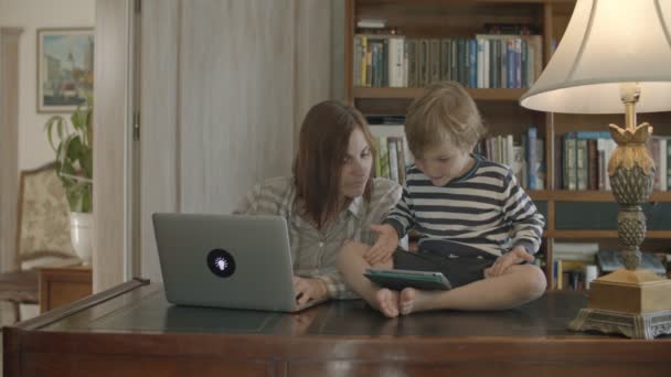 Блондин мальчик тревожит работающую мать, играющую на планшете сидя на деревянном столе — стоковое видео