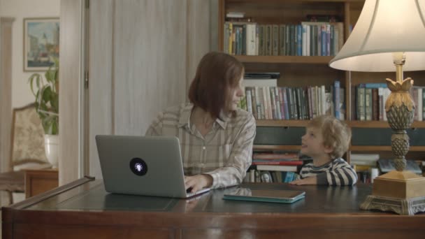 Блондин мальчик тревожит работающую мать, играющую на планшете сидя на деревянном столе — стоковое видео
