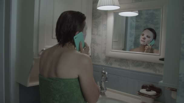 Slow motion of brunette female in towel in cosmetic mask on her face with smartphone after shower — Stock Video