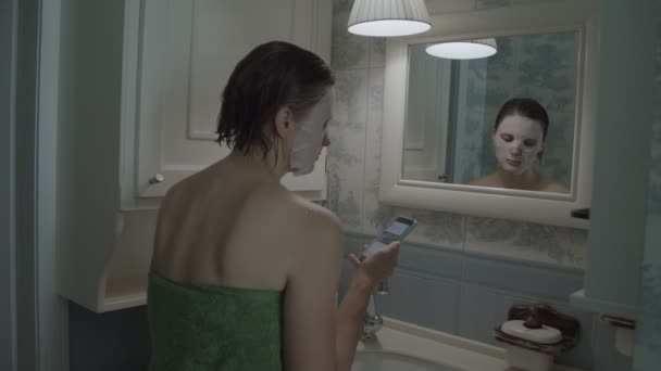 Cámara lenta de morena hembra en toalla en máscara cosmética en su cara con teléfono inteligente después de la ducha — Vídeos de Stock