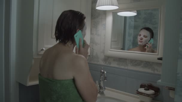 シャワー後にスマートフォンで彼女の顔に化粧品マスクでタオルでブルネットの女性のスローモーション — ストック動画