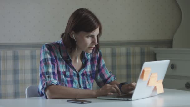 Giovane freelance donna che lavora su laptop e smartphone con auricolari seduti al tavolo bianco in posizione di provenienza — Video Stock