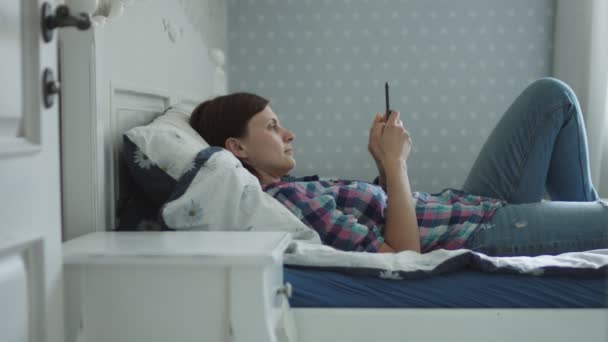 Attraente giovane donna sdraiata sul letto e navigare online tramite smartphone e laptop — Video Stock