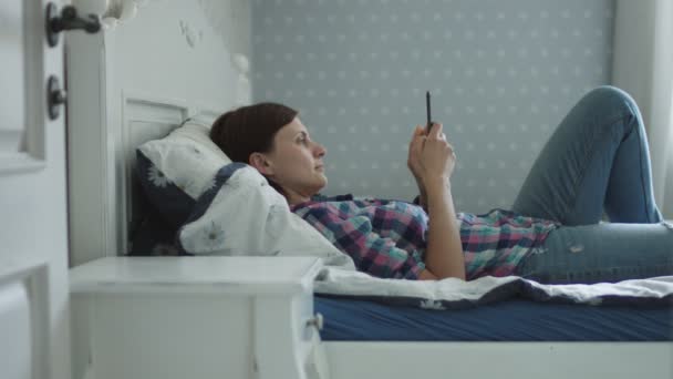 Attractive young female lying on the bed and surfing online via smartphone and laptop — Stock Video