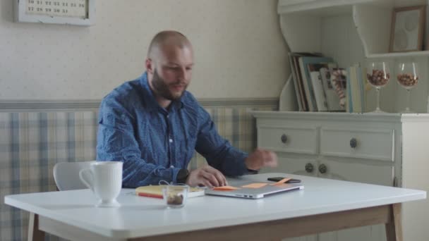 Freelancer masculino jovem trabalhando em laptop e smartphone sentado à mesa com chá e lanches — Vídeo de Stock