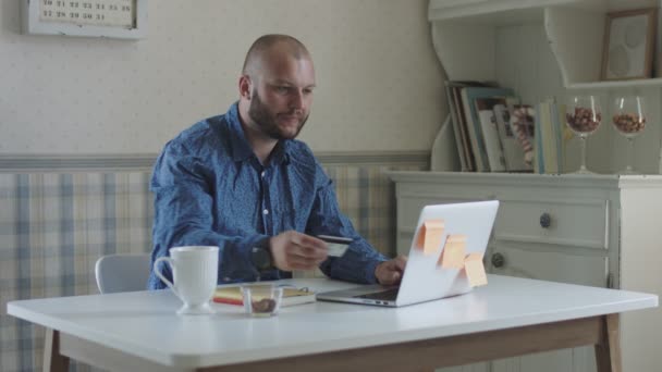 Freelancer masculino jovem fazendo compra via cartão de crédito e trabalhando no laptop sentado à mesa com chá e lanches — Vídeo de Stock