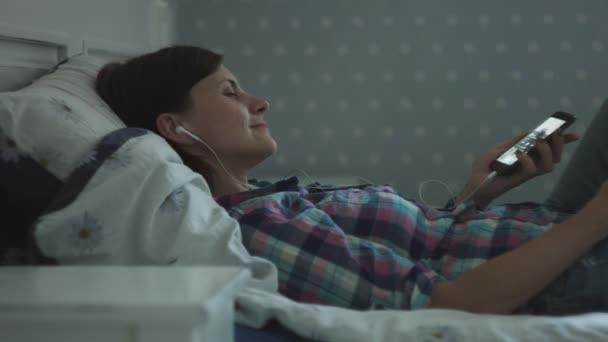 Close up of attractive young female lying on the bed and listening the music in earphones. Women relaxing by enjoying the music — Stock Video