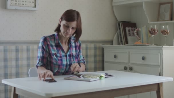 若い女性がレシピ本を通して親指を立て、明るいインテリアでテーブルの上に座ってスマートフォンで写真を撮る. — ストック動画