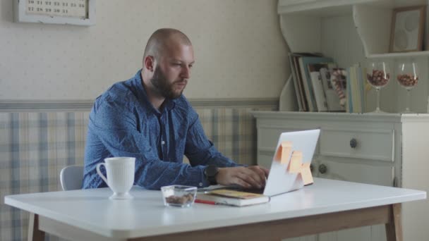 Joven hombre freelance trabajando en el ordenador portátil y teléfono inteligente sentado a la mesa con té y aperitivos — Vídeo de stock