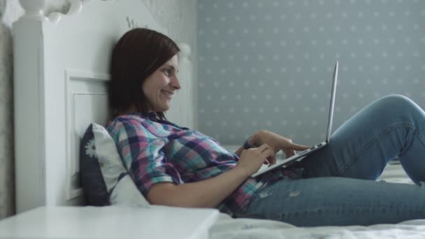 魅力的な若い女性がベッドに横たわり、スマートフォンやラップトップを介してオンラインサーフィン — ストック動画