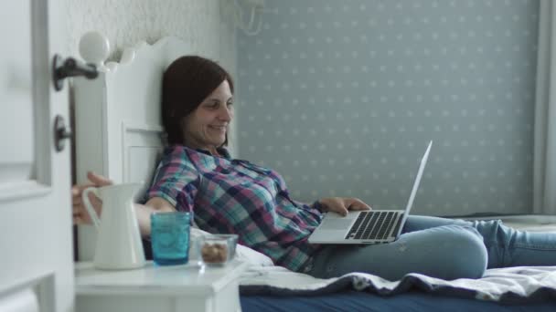 Приваблива молода жінка лежить на ліжку і серфінг онлайн через смартфон і ноутбук — стокове відео