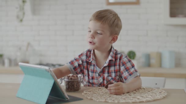 Carino ragazzo bambino biondo mangiare snack senza glutine con latte vegan e guardare cartoni animati su tablet in cucina luminosa al rallentatore — Video Stock