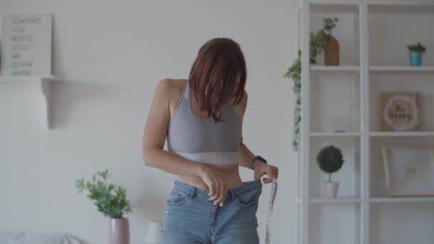Молода жінка захоплюється результатом втрати ваги під час носіння старих джинсів у спальні. Щаслива жінка фотографує живіт після того, як ви схудли . — стокове відео