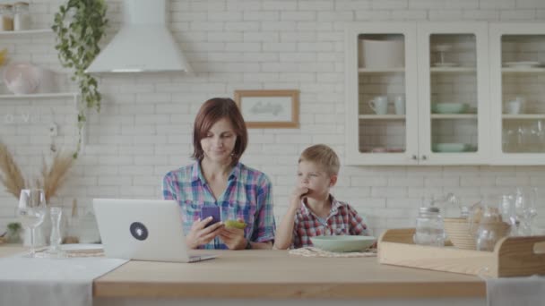 Madre trabajando en el ordenador portátil con niño comiendo galletas de chocolate y bebiendo yogur en la cocina blanca — Vídeos de Stock