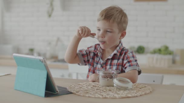 Lindo niño rubio comiendo bocadillos sin gluten con leche vegana y viendo dibujos animados en la tableta en la cocina brillante en cámara lenta — Vídeos de Stock