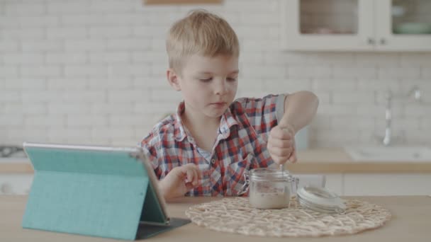 Милий блондинка малюк їсть закуски без глютену з веганським молоком і дивиться мультфільми на планшеті на яскравій кухні в повільному русі — стокове відео