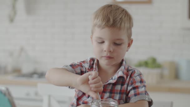 かわいいブロンドの幼児の男の子は、ビーガンミルクとグルテンフリーのスナックを食べて、スローモーションで明るいキッチンでタブレットで漫画を見て — ストック動画