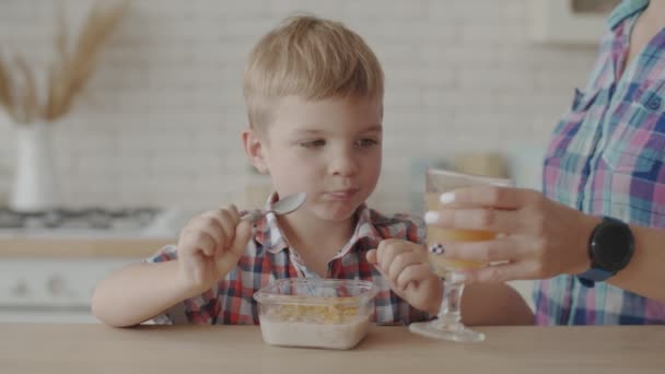 Fröhlicher kleiner Junge, der glutenfreie Cornflakes mit veganer Milch isst und Apfelsaft trinkt. Mutter hilft ihrem Sohn beim Frühstück — Stockvideo