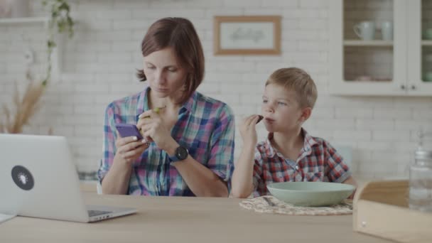 Madre trabajando en el ordenador portátil con niño comiendo galletas de chocolate y bebiendo yogur en la cocina blanca — Vídeos de Stock