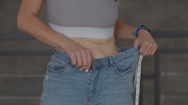 Ung kvinna beundrar resultatet av viktminskning medan du bär gamla jeans i sovrummet. Lycklig kvinna tar bild av hennes mage efter att du gått förlorad vikt. — Stockvideo