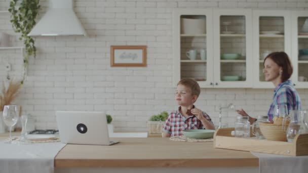 Anya dolgozik-ra laptop-val totyogó fiú étkezési csokoládé süti és ivás joghurt-ra fehér konyha — Stock videók