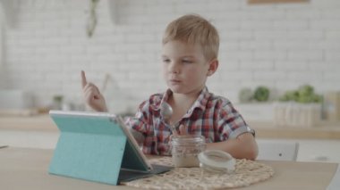Sevimli sarışın yürümeye başlayan çocuk vegan süt ile glutensiz aperatifler yeme ve yavaş hareket parlak mutfakta tablet karikatürler izlerken