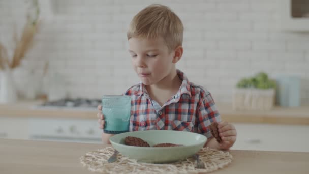 Хлопчик Тоддлер їсть шоколадне печиво і п'є йогурт, сидячи на яскравій кухні сам у повільному русі — стокове відео