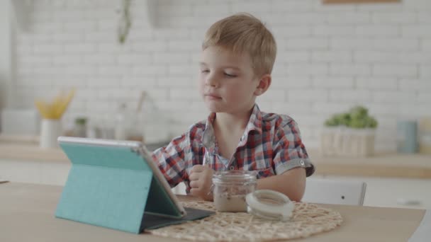 Милый блондин-мальчик ест безглютеновые закуски с веганским молоком и смотрит мультики на планшете на яркой кухне в замедленной съемке — стоковое видео