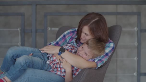 Счастливая молодая брюнетка мать играет с блондинкой ребенок сын сидит в кресле. Женщина и мальчик смеются в замедленной съемке . — стоковое видео