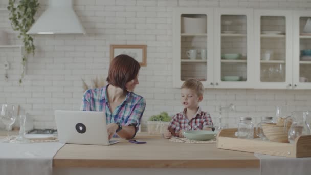 Mutter arbeitet am Laptop, Kleinkind isst Schokokekse und trinkt Joghurt auf weißer Küche — Stockvideo