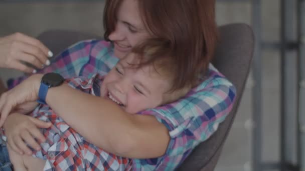 Joyeux jeune mère brune jouant avec fils blond tout-petit assis dans un fauteuil. Femme et garçon riant au ralenti . — Video