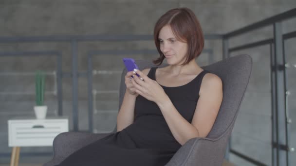Belle giovani donne è felice di chattare in smartphone seduto su un accogliente divano. Attraente femmina in abito nero facendo selfie sul cellulare — Video Stock