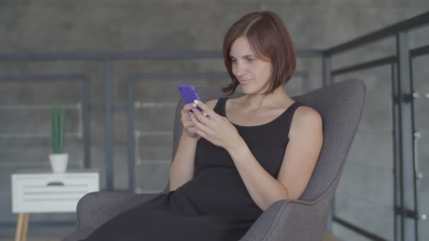 Bonitas mujeres jóvenes está feliz con el chat en el teléfono inteligente sentado en el sofá acogedor. Atractiva hembra en vestido negro haciendo selfie en el teléfono celular — Vídeo de stock