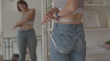 Yatak odasında eski kot pantolon giyerken kilo vermenin sonuçlarına hayran olan genç bir kadın. Kilo verdiğinde karnının fotoğrafını çeken mutlu bir kadın.. 