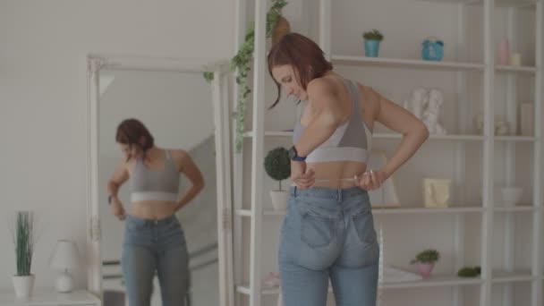 寝室の古いジーンズを身に着けている間若い女性は減量の結果を賞賛する。幸せ女性撮影彼女の腹のとき彼女失われた重量. — ストック動画