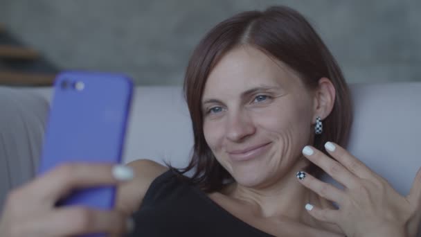 Όμορφες νέες γυναίκες είναι ευχαριστημένοι με τη συνομιλία στο smartphone κάθεται σε άνετο καναπέ. Ελκυστική γυναίκα με μαύρο φόρεμα κάνοντας selfie στο κινητό τηλέφωνο — Αρχείο Βίντεο