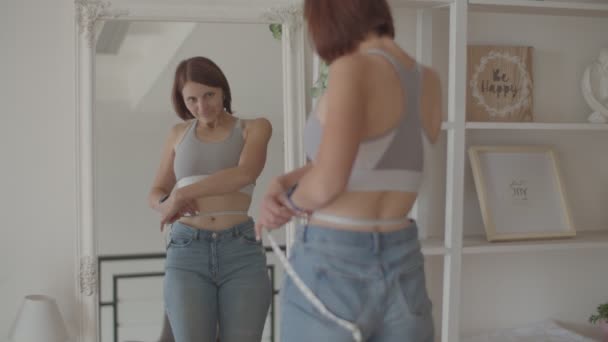 Молодая женщина любуется результатом потери веса во время ношения старых джинсов в спальне. Счастливая женщина фотографировала свой живот, когда похудела. . — стоковое видео