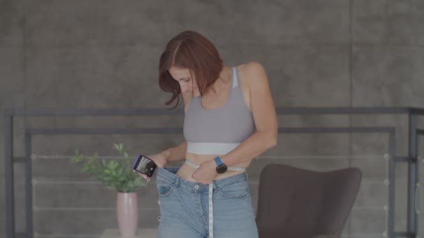 Jonge vrouw bewondert het resultaat van gewichtsverlies tijdens het dragen van oude jeans in de slaapkamer. gelukkig wijfje het nemen van foto van haar buik toen ze afviel. — Stockvideo
