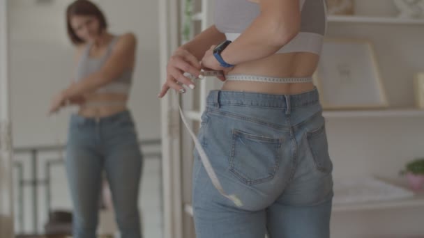 Молода жінка захоплюється результатом втрати ваги під час носіння старих джинсів у спальні. Щаслива жінка фотографує живіт, коли вона схудла . — стокове відео