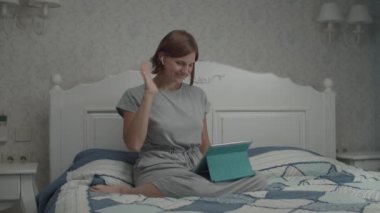 Gri elbiseli, kablosuz kulaklıklar ve tablet bilgisayarla internetten konuşan genç ve güzel kadınların yavaş çekimleri..