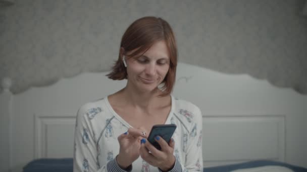 Slow motion van jonge mooie vrouwen in pyjama surfen op internet met haar smartphone en draadloze oortelefoons zitten op het bed. — Stockvideo