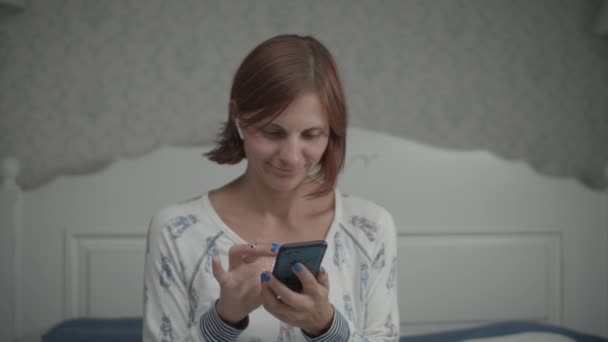 Slow motion van jonge mooie vrouwen in pyjama surfen op internet met haar smartphone en draadloze oortelefoons zitten op het bed. — Stockvideo
